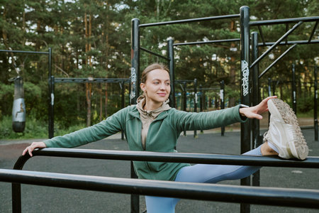 Young adult woman stretching leg on outdoor fitness equipment in park, smiling and looking away from...