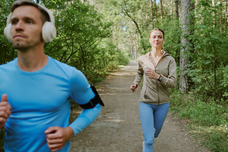 Caucasian young adult man running with headphones in foreground while Caucasian young adult woman...