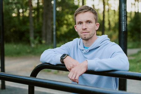 Portrait of young adult man standing outdoors leaning on metal railing looking into camera with...