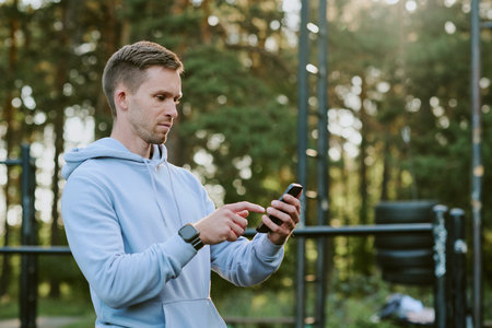 Caucasian young adult man standing outdoors using smartphone with focused expression, holding device in both hands, wearing smartwatch, fitness equipment visible in backgroundの写真素材