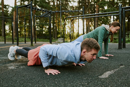 Young adult man and young adult woman exercising outdoors performing push ups on sports ground, both...