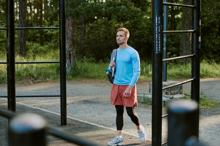 Caucasian young adult man walking outdoors in park carrying sports bag over shoulder passing outdoor...