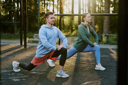 Caucasian young adult man and Caucasian young adult woman performing lunges outdoors in park...