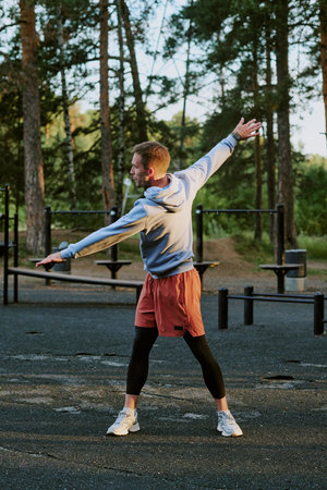 Young adult man stretching arms outdoors in park, standing on asphalt surface with exercise equipment in background, wearing athletic clothing, facing away from cameraの写真素材