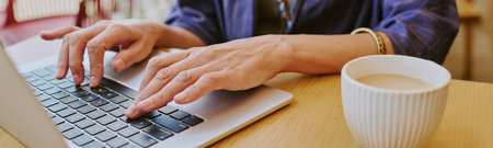 Website banner of woman typing on laptop keyboard with both hands, working at table with cup of...