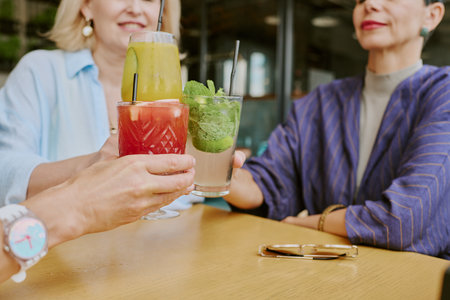 Three middle aged Caucasian women sitting at table clinking colorful cocktails, smiling and enjoying social gathering, hands and drinks in focus, faces partially visibleの写真素材
