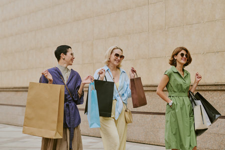 Three middle aged Caucasian women walking outdoors carrying shopping bags, smiling and talking together, wearing sunglasses, enjoying leisure time, urban background visibleの写真素材