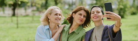Website banner of three middle aged Caucasian women smiling and posing for selfie outdoors in park,...