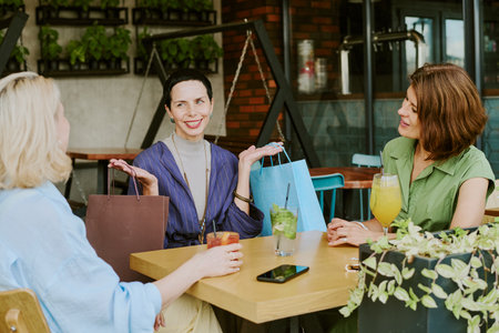 Three young adult Caucasian women sitting at outdoor cafe table smiling and talking while holding shopping bags and drinks, engaging in friendly conversation during daytimeの写真素材