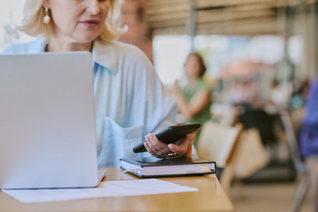 Middle aged woman using smartphone while sitting at desk with laptop and notebook, focusing on...