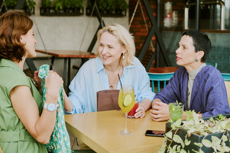 Three middle aged Caucasian women sitting at table talking and smiling, one woman holding patterned fabric, drinks with lime and mint on table, outdoor cafe settingの写真素材