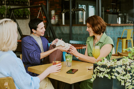 Three middle aged Caucasian women sitting at table in cafe, smiling and exchanging gift, drinks and...