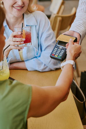 Caucasian middle aged woman holding drink while making contactless payment with credit card on point of sale terminal, another woman with beverage sitting nearby, hands and faces visibleの写真素材