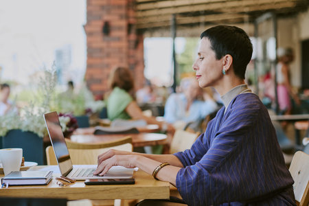 Middle aged Caucasian woman working on laptop at outdoor cafe, sitting at table with notebook and smartphone, focusing on screen, short dark hair, side profile visibleの写真素材