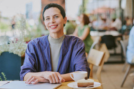 Portrait of middle aged Caucasian woman smiling at camera while sitting at table with laptop, coffee cup and dessert in modern cafe, short dark hair visibleの写真素材