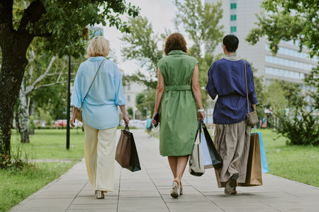 Three middle aged women walking outdoors on city sidewalk carrying shopping bags, backs facing...