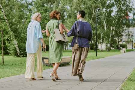 Three adult women walking together on park pathway, engaging in conversation, carrying handbags, surrounded by green trees and grassの写真素材