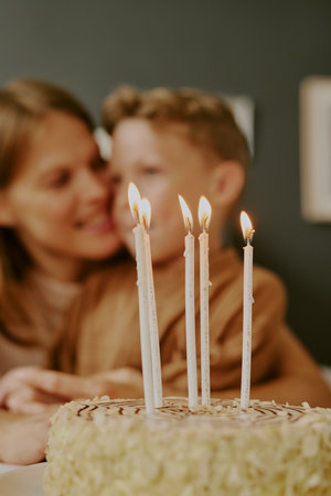 Caucasian woman embracing Caucasian boy in background, both smiling and looking at each other, birthday cake with five lit candles in foreground, celebrating special occasionの写真素材