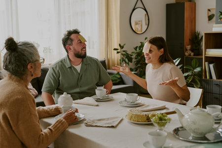 Caucasian senior woman, Caucasian middle aged man, Caucasian middle aged woman sitting at dining table playing guessing game with sticky notes on foreheads, smiling and gesturingの写真素材