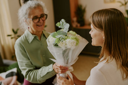 Senior Caucasian woman smiling while receiving bouquet of flowers from young adult Caucasian woman indoors, both women interacting closely and expressing positive emotionsの写真素材
