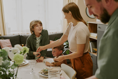 woman serving food to two boys at dining table while middle aged man standing nearby, older boy smiling and younger boy sitting, family gathering at homeの写真素材