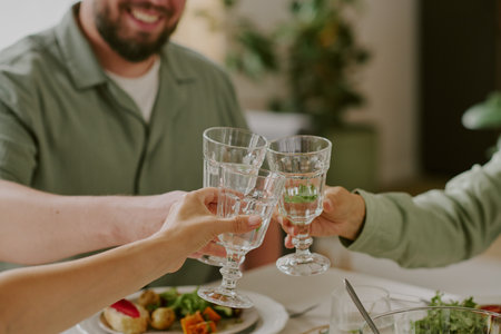 Three young adults clinking glasses while sitting at table, celebrating together, hands and partial...