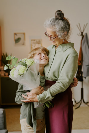 Caucasian senior woman embracing Caucasian boy while holding bouquet of flowers, both smiling and making eye contact, standing together in home setting, showing affectionate momentの写真素材