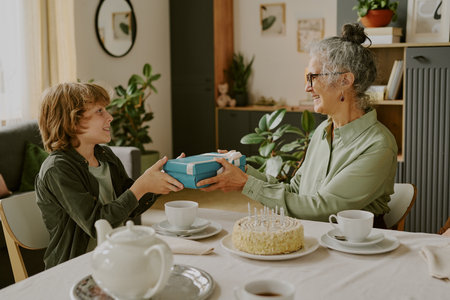 Caucasian senior woman giving wrapped gift to boy while sitting at table with birthday cake and tea set, both smiling and making eye contact in cozy home settingの写真素材
