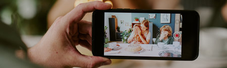 Website banner of Caucasian woman with two Caucasian children sitting at table celebrating birthday, smartphone recording family moment at homeの写真素材