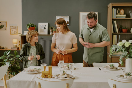 Caucasian boy talking with woman and man standing together near dining table set for meal, all smiling and interacting in modern home dining roomの写真素材