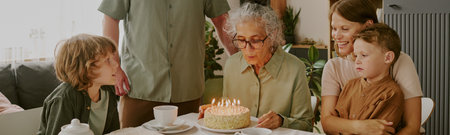 Website banner of Senior Caucasian woman blowing out candles on birthday cake surrounded by middle aged Caucasian woman, young Caucasian boy, young Caucasian man and child celebrating togetherの写真素材