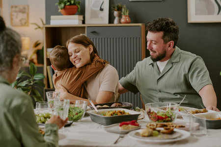 Caucasian young woman hugging child while sitting at dining table with middle aged Caucasian man and senior woman, sharing family meal and enjoying togetherness during gatheringの写真素材