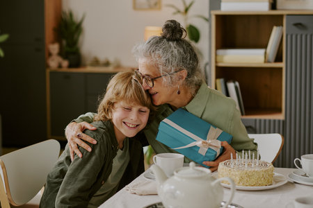 Senior Caucasian woman hugging smiling Caucasian boy while giving wrapped gift at table with birthday cake and candles, both sitting close together celebrating special occasionの写真素材