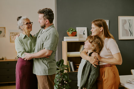Middle aged Caucasian man embracing senior Caucasian woman while young Caucasian woman hugging Caucasian boy, all standing together in modern home interior smiling warmlyの写真素材