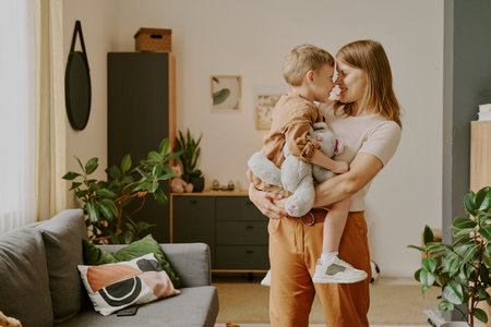 Caucasian young adult woman holding Caucasian child boy with stuffed animal, standing close together and smiling in modern living room, plants and decor visible in backgroundの写真素材