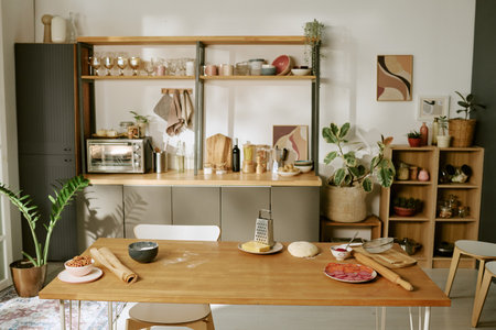 Wooden kitchen table holding rolling pin, dough, grated cheese, sliced sausage, and bowl of beans,...