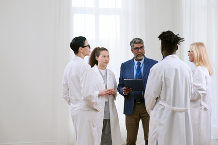 Middle aged Caucasian man with gray beard holding clipboard, talking to diverse group of young adult doctors including Black man and Caucasian women standing in medical settingの写真素材