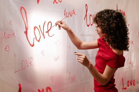 Young adult Caucasian woman painting word love and drawing hearts on transparent wall, celebrating Valentines Day, standing in profile with curly hair, wearing elegant dressの写真素材