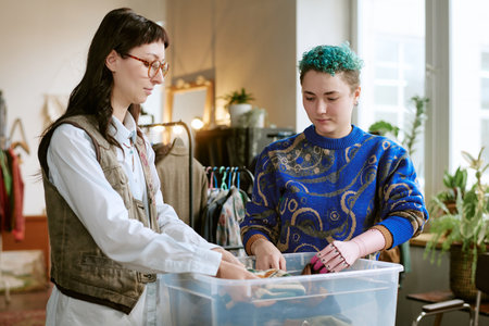 Young woman with prosthetic hand sorting second hand clothing with her colleague in transparent...