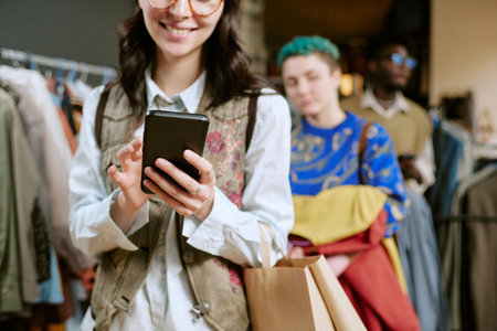 Young adult woman smiling while using smartphone and holding shopping bag in second hand clothing...