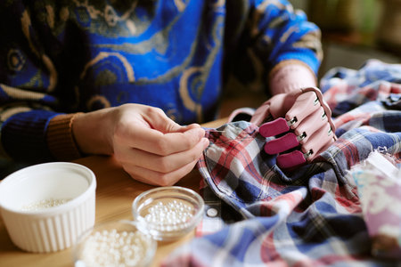 Caucasian young adult woman with prosthetic hand repairing second hand plaid shirt using needle and thread, holding fabric while sewing, glass bowls with buttons on wooden tableの写真素材