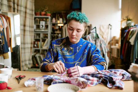 Young adult Caucasian woman with prosthetic hand repairing second hand clothing at table, using...