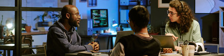 Three young adult professionals collaborating at desk in modern office, Black man speaking while man...