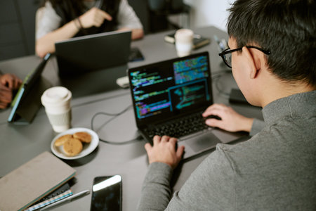 Caucasian young adult man working on laptop with code on screen, sitting at desk with coffee and...