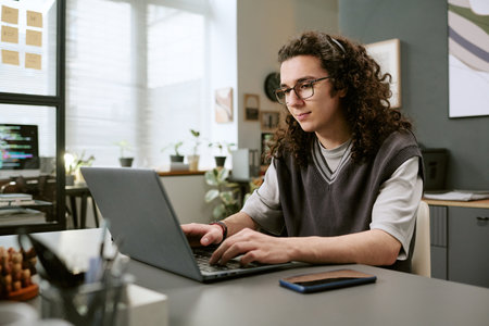 Young adult Caucasian man with long curly hair sitting at desk working on laptop computer in modern...