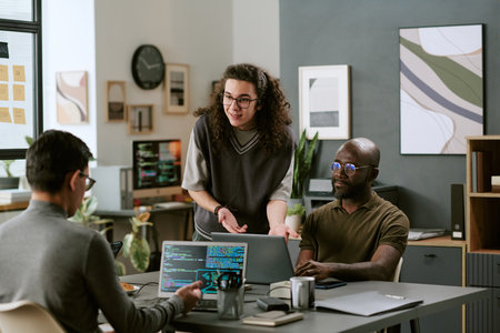 Young adult Caucasian man standing and gesturing while talking to young adult Caucasian man and young adult Black man, sitting at desk using laptops in modern office workspaceの写真素材
