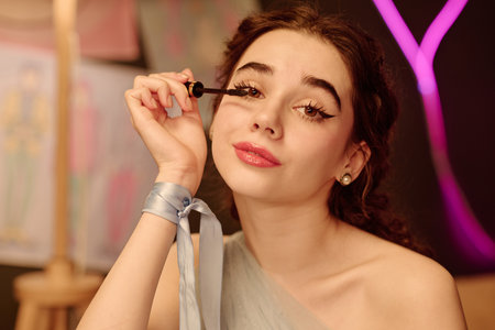 Portrait of Caucasian teenage girl applying mascara to eyelashes, looking directly at camera with relaxed expression, sitting indoors with blurred colorful background artworkの写真素材