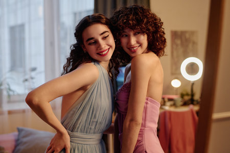 Two young adult women smiling and posing together in elegant dresses, standing close to each other in indoor setting, looking happy and confidentの写真素材