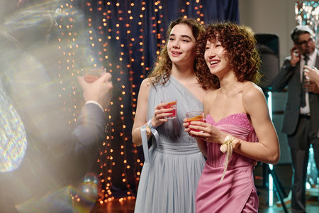 Teen girls standing close together smiling and holding drinks at festive party, looking toward in foreground holding glassの写真素材