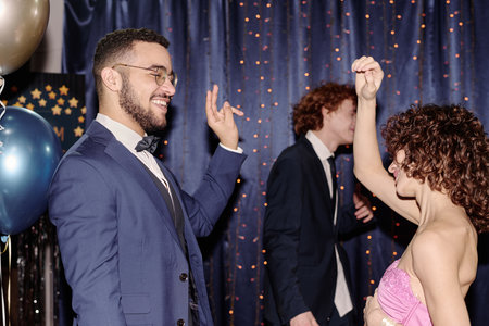 Young adult man smiling and dancing with young adult Caucasian woman raising arm at party, another young adult Caucasian man walking in background, festive setting with balloons and lightsの写真素材